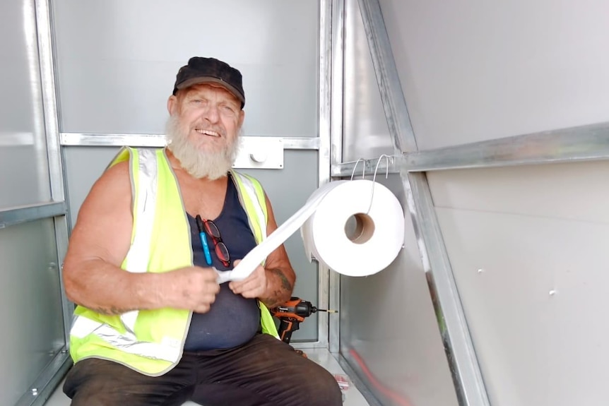 A smiling bearded man in a high vis jacket sitting in a toilet cubicle next to a drill holding a roll of toilet paper