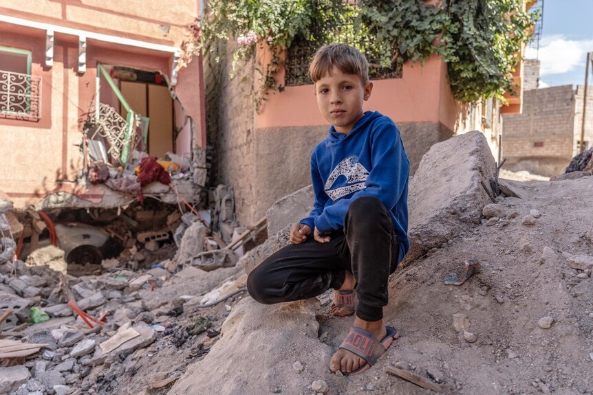 A young boy looking at the camera while kneeling near a pile of rubble.