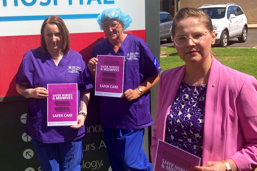 Three women, two of them nurses, stand in front of a hospital sign holding petition forms 
