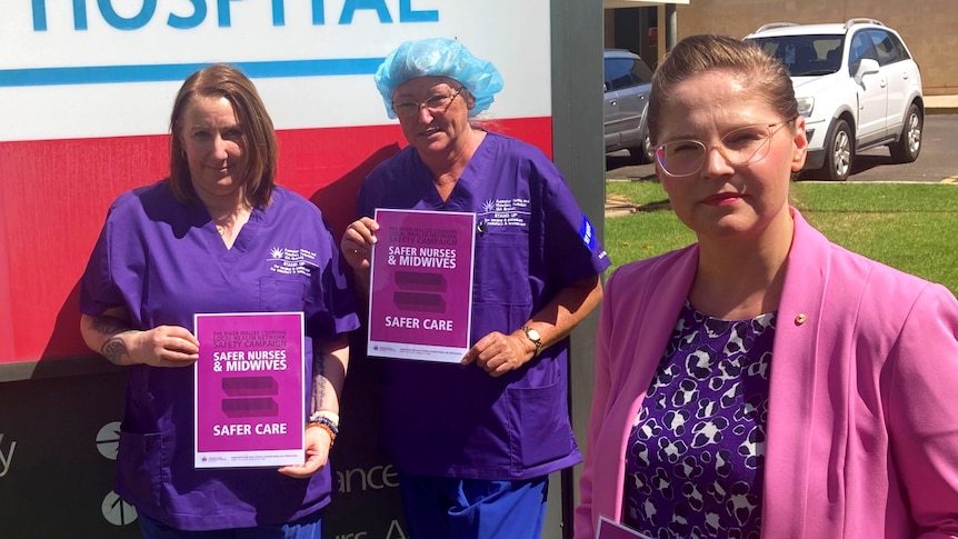 Three women, two of them nurses, stand in front of a hospital sign holding petition forms 