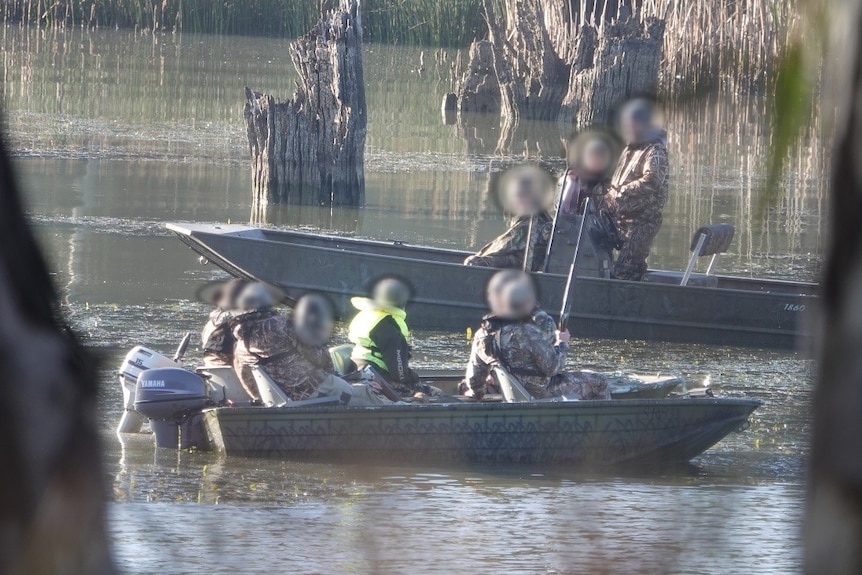 A group of hunters in khaki travel down a river in tin boats.