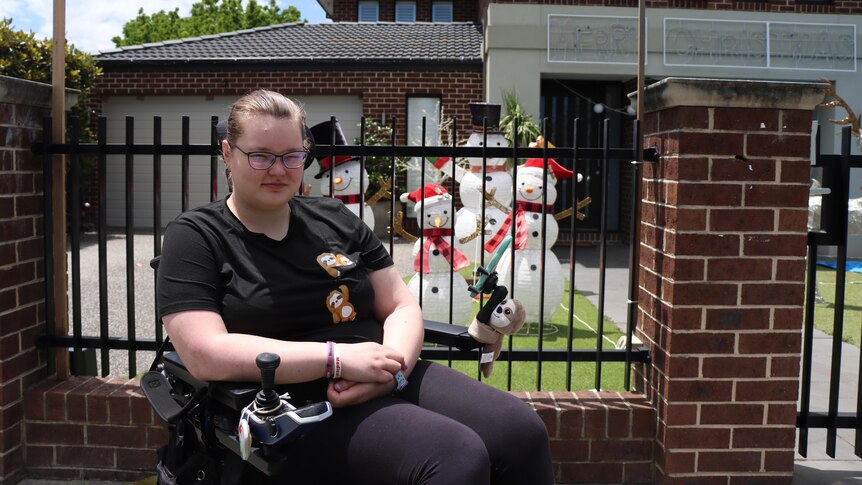 Jessi Hooper, wearing all black, sits in her wheelchair in front of a home with Christmas decorations.