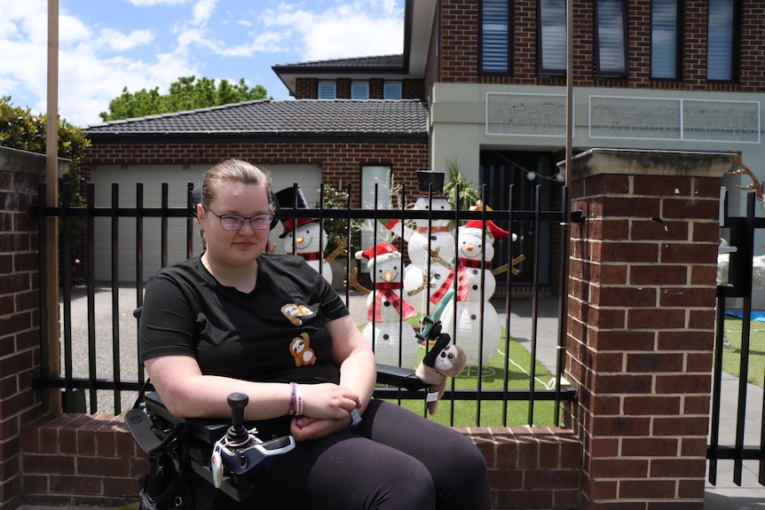 Jessi Hooper, wearing all black, sits in her wheelchair in front of a home with Christmas decorations.