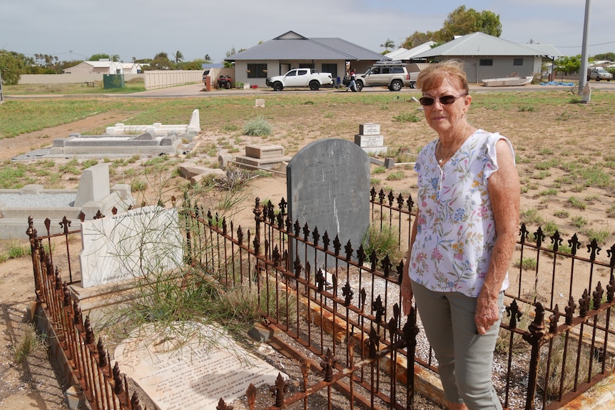 A stern woman in her sixties stands in front of a grave with a desert suburban backdrop. The grave is snapped in two.