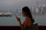 A woman in a mask looks at her smartphone while standing on a ferry, with the sea and the city of Hong Kong in the background.