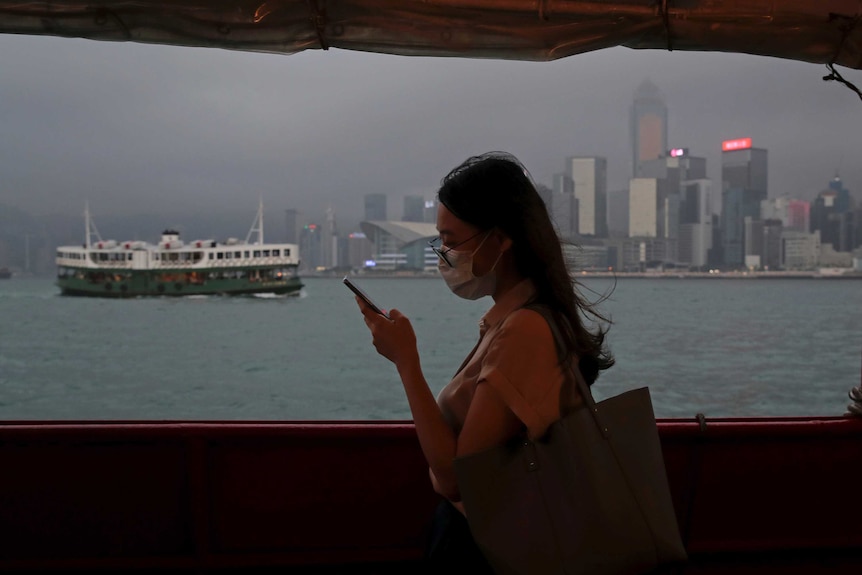 A woman in a mask looks at her smartphone while standing on a ferry, with the sea and the city of Hong Kong in the background.