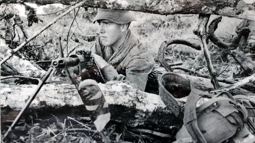 Alan Wilkinson lies down resting his arm on a machine gun around trees.