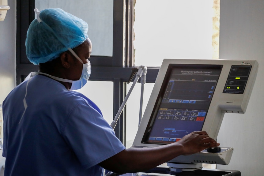 A nurse in scrubs, a hair net and face mask stands next to a machine in a hospital ward.