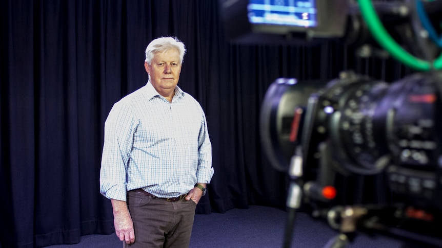 A man with white hair stands in a television studio in front of a camera