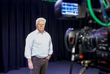 A man with white hair stands in a television studio in front of a camera