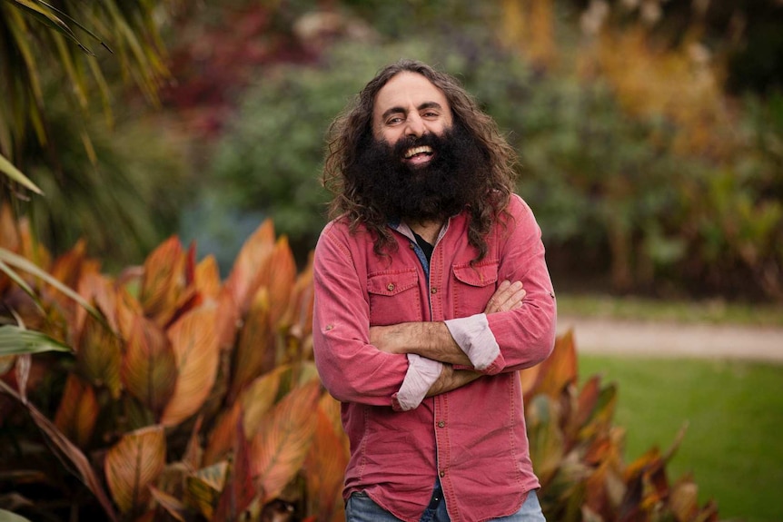 Man with beard and long dark hair standing cross armed in a garden. 