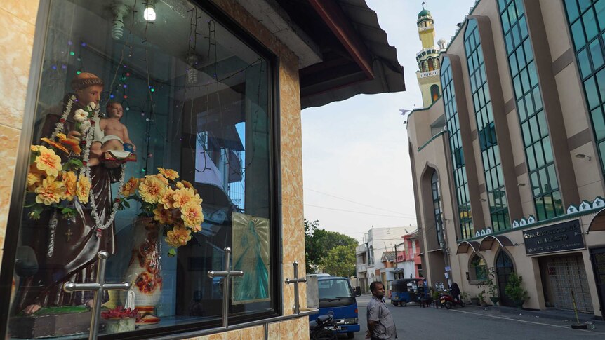 Yellow flowers in vases and a statue of the Virgin Mary in a window with a streetscape in the background.