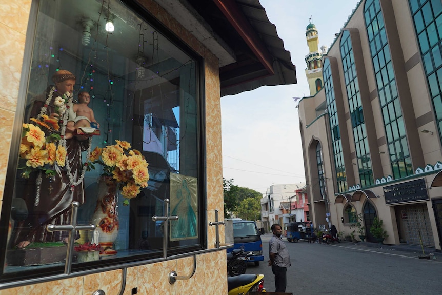 Yellow flowers in vases and a statue of the Virgin Mary in a window with a streetscape in the background.