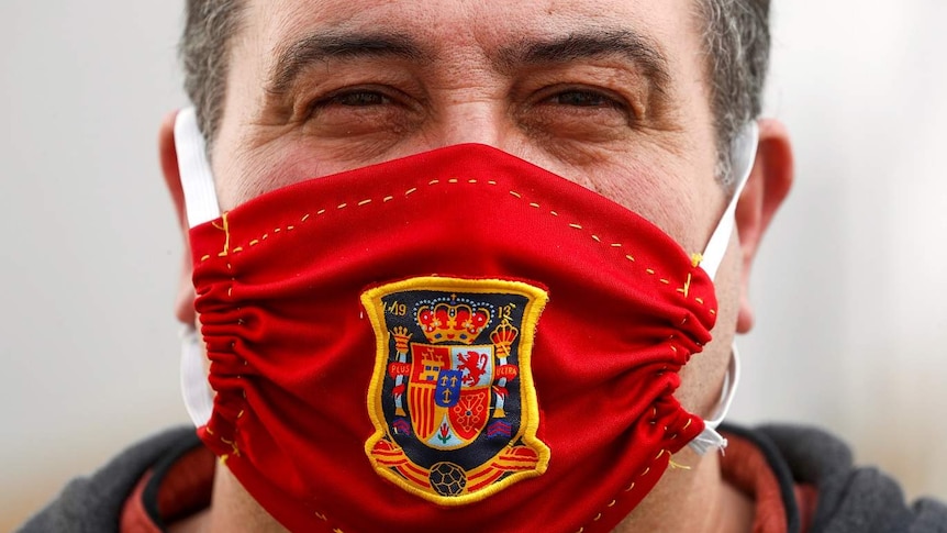 A man wears a red face mask with a yellow and black coat of arms on it.