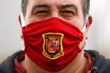A man wears a red face mask with a yellow and black coat of arms on it.