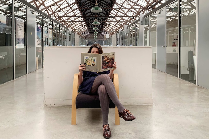 Mia Timpano sitting cross-legged on a chair and reading an arts book, which partially covers her face.