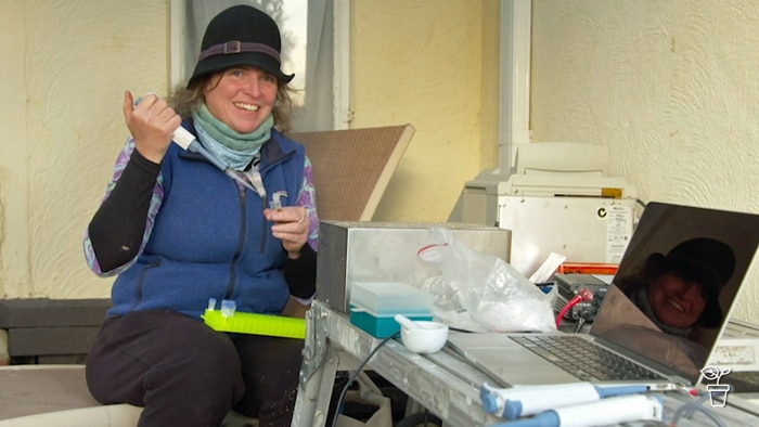 Woman in a hat sitting amongst laboratory equipment and using a pipette to fill a container