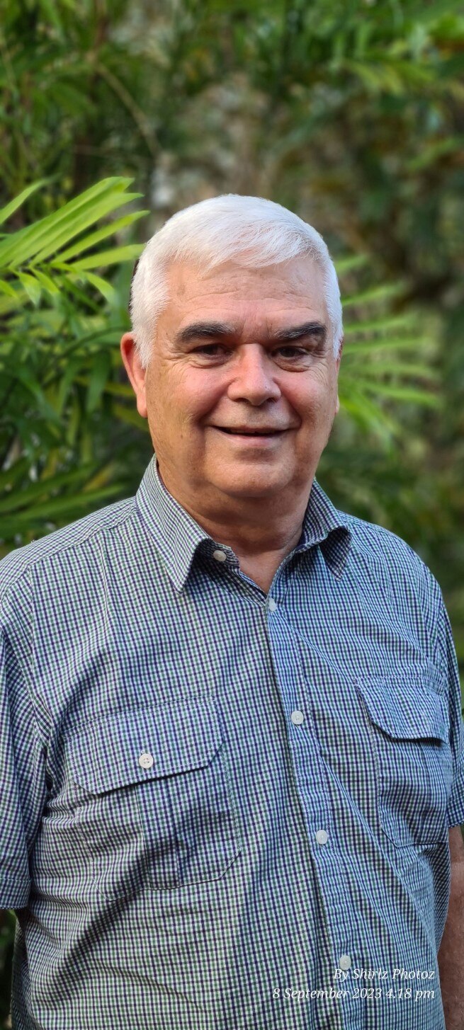 An older, grey-haired man stands in front of some greenery.