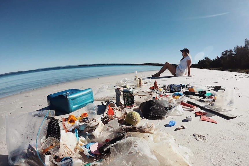 A pile of rubbish on a white sand beach, with a woman sitting nearby.