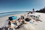 A pile of rubbish on a white sand beach, with a woman sitting nearby.