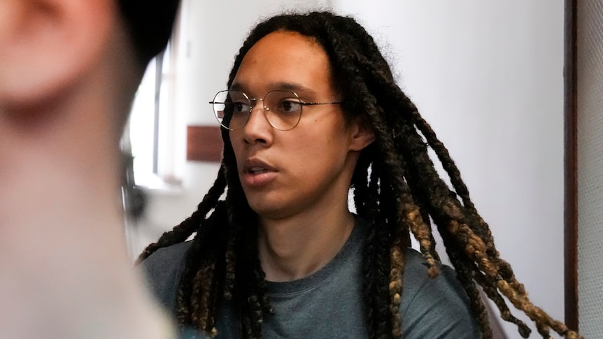 A dark-skinned woman with long, dark dreadlocks and round, gold-rimmed glasses is wearing a prison T-shirt