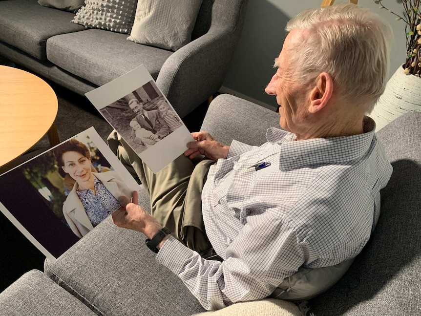A man sitting on a couch looks down, smiling, at photos of his mother and father.