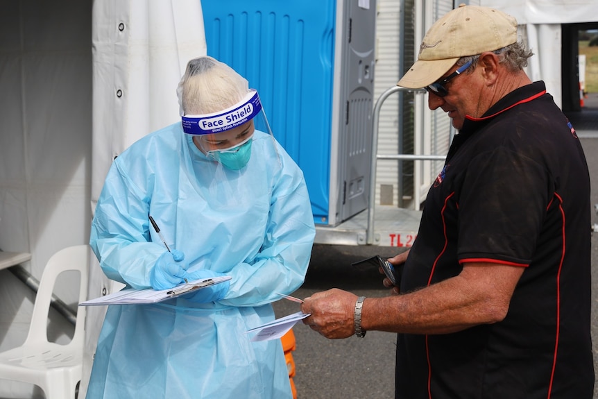 A woman nurse in full PPE filling out paperwork on a clipboard, looking at a card that a man is holding out to her