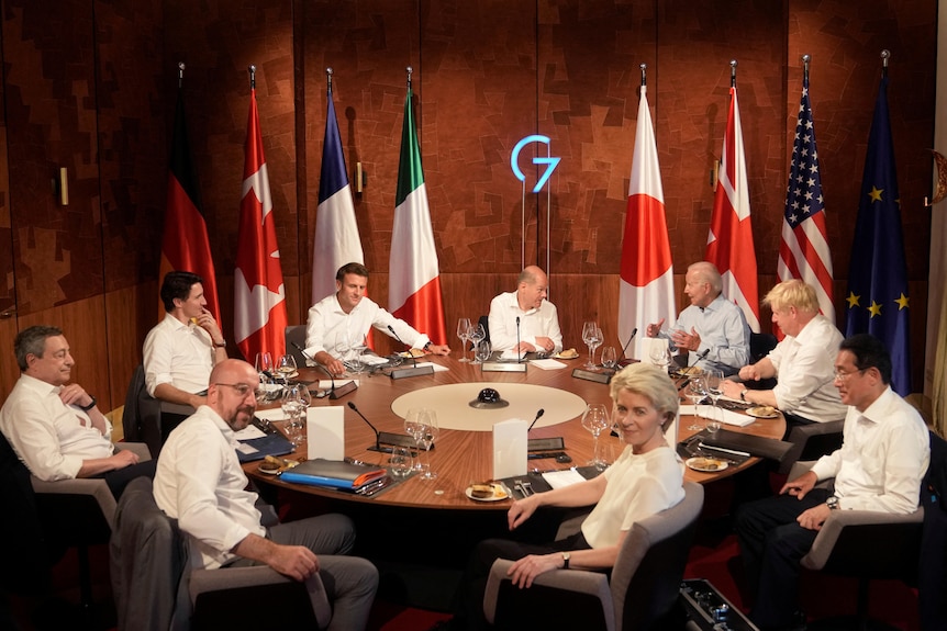 G7 leaders sitting around round table with respective flags lined up against wall.