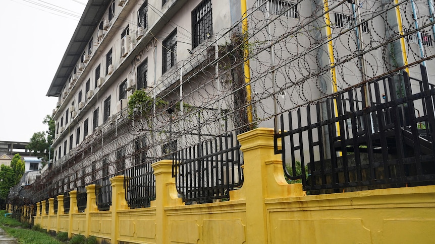A grey building surrounded by yellow brick and black iron barred fences with barbed wire on top