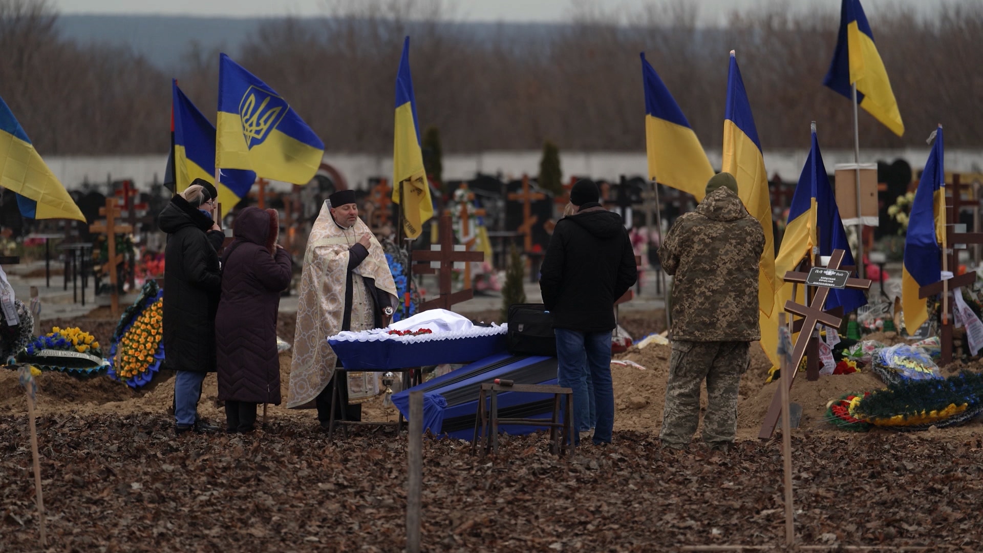 Men and women and a priest stand around an open coffin in a cemetery. They are surrounded by Ukrainian flags.