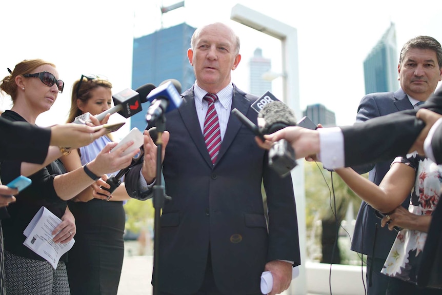 John Day speaks to a group of reporters at Elizabeth Quay, with Perth's CBD in the background.