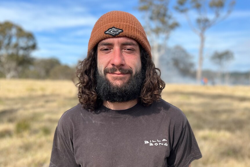 Young man with dark curly hair and a beard, wearing an orange beanie, stands in paddock smiling.