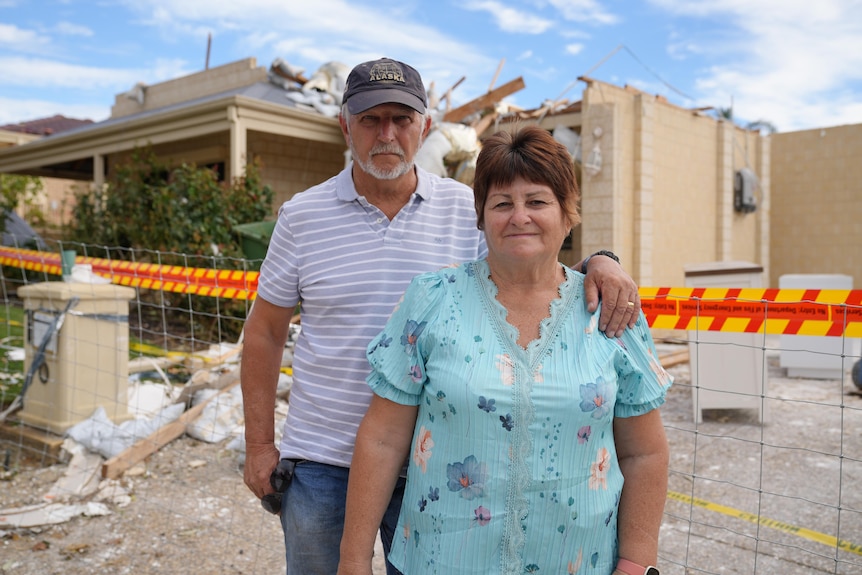 A man and woman standing in front of their wrecked home, which has been taped off.