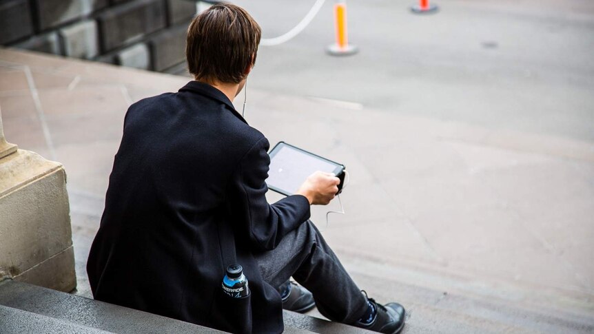 A boy in a school uniform sits on the steps of State Parliament looking at a tablet.