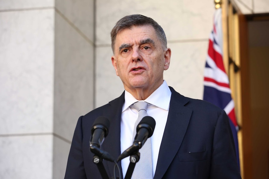 Brendan Murphy stands at a podium in a courtyard with an Australian flag behind him