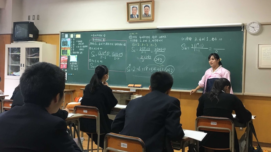 A teacher wearing a pink traditional dress from North Korea stands in front of a blackboard filled with algebra.