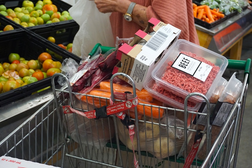 A close-up shot of a shopping trolley full of groceries in a supermarket with a woman behind bagging apples.