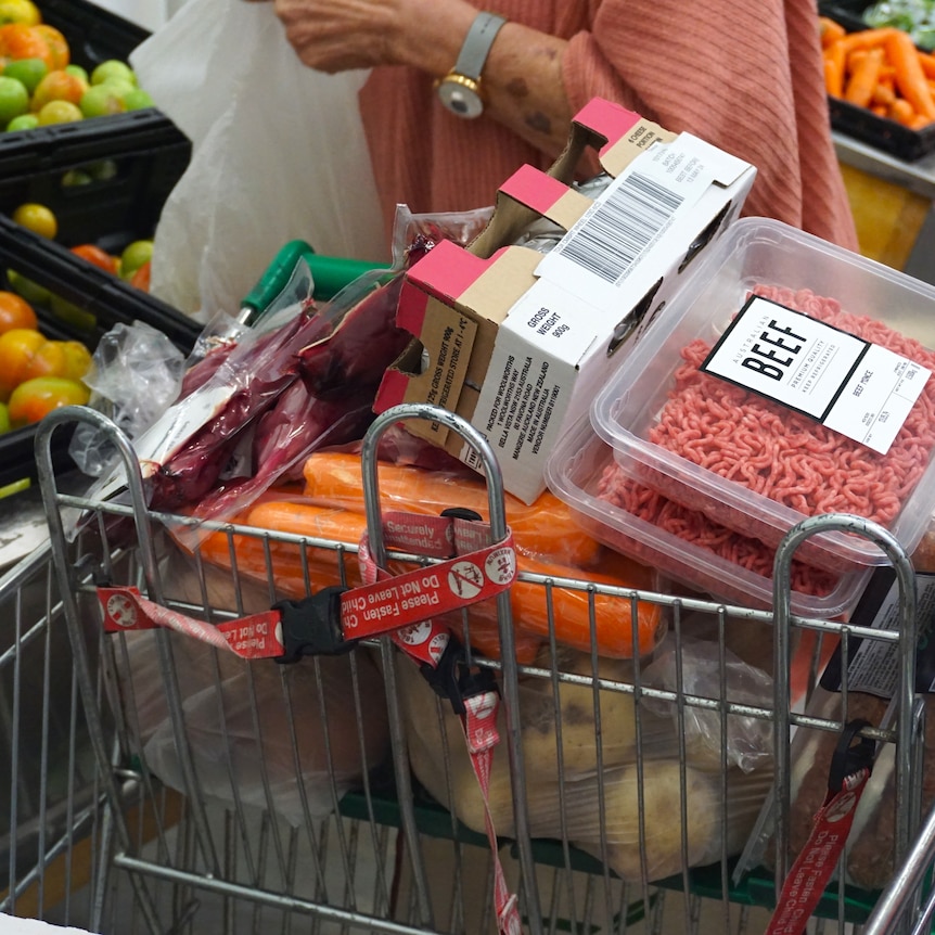 A close-up shot of a shopping trolley full of groceries in a supermarket with a woman behind bagging apples.