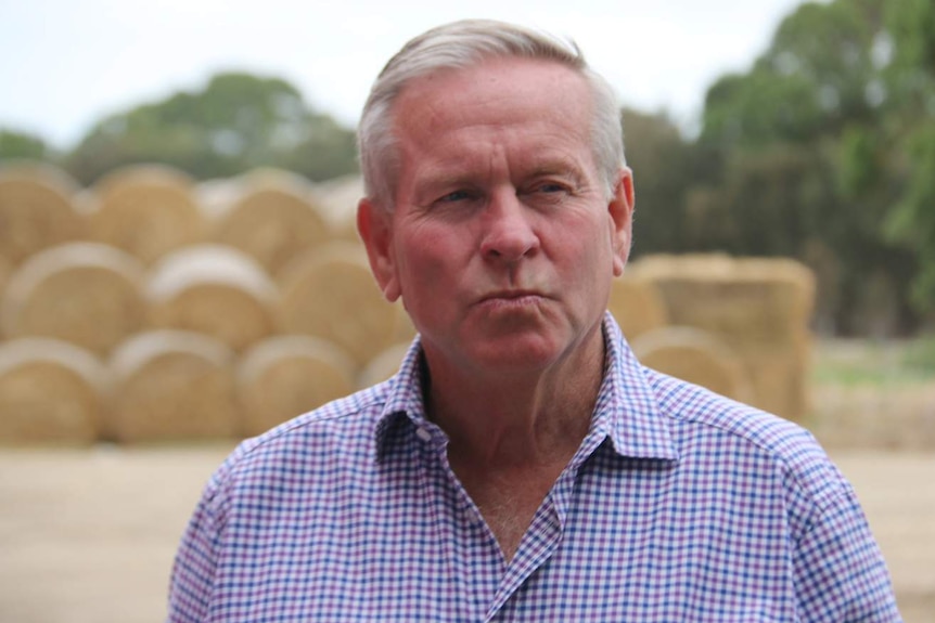 A head and shoulders shot of Colin Barnett with his lips pursed, standing in front of hay bales.