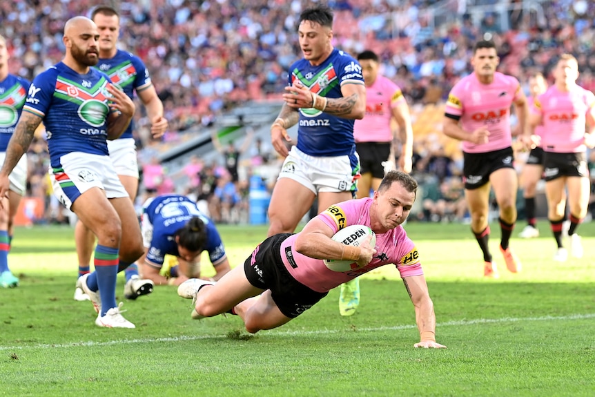A Penrith NRL player in a pink jersey dives to the ground while holding the ball to score a try while opposition players watch.