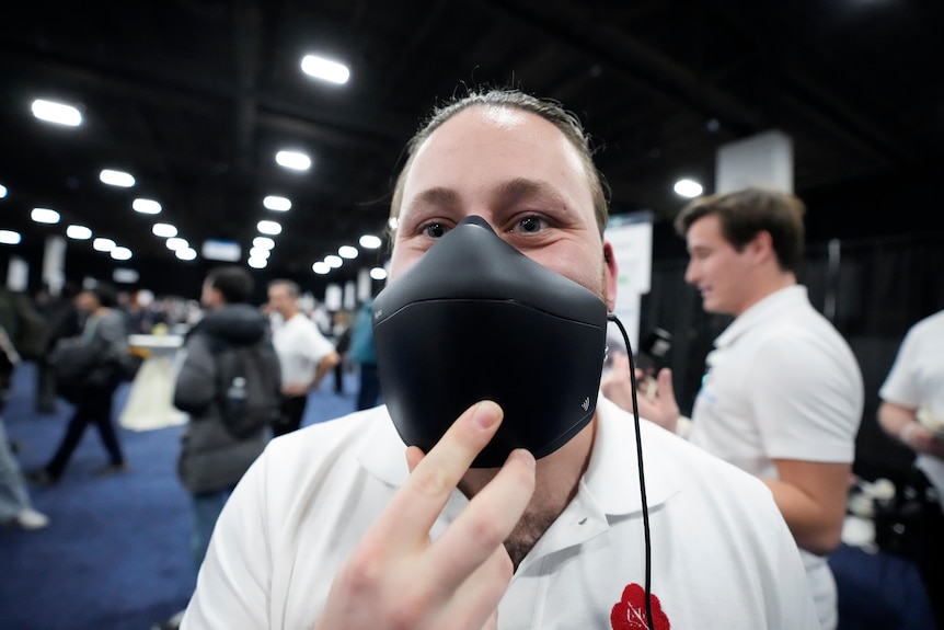 A close up of a man holding a thick face mask over his mouth with one hand, indoors at a technology industry event