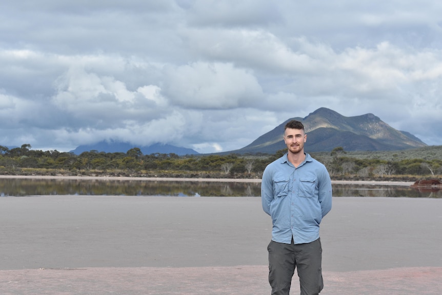 Man standing in front of salt lake wearing blue shirt and grey pants with mountain in the background