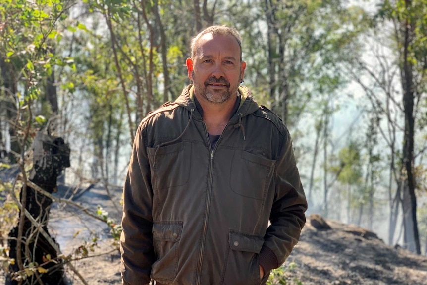 Man wearing a brown jacket standing in front of smoking bushland which has just been burnt.