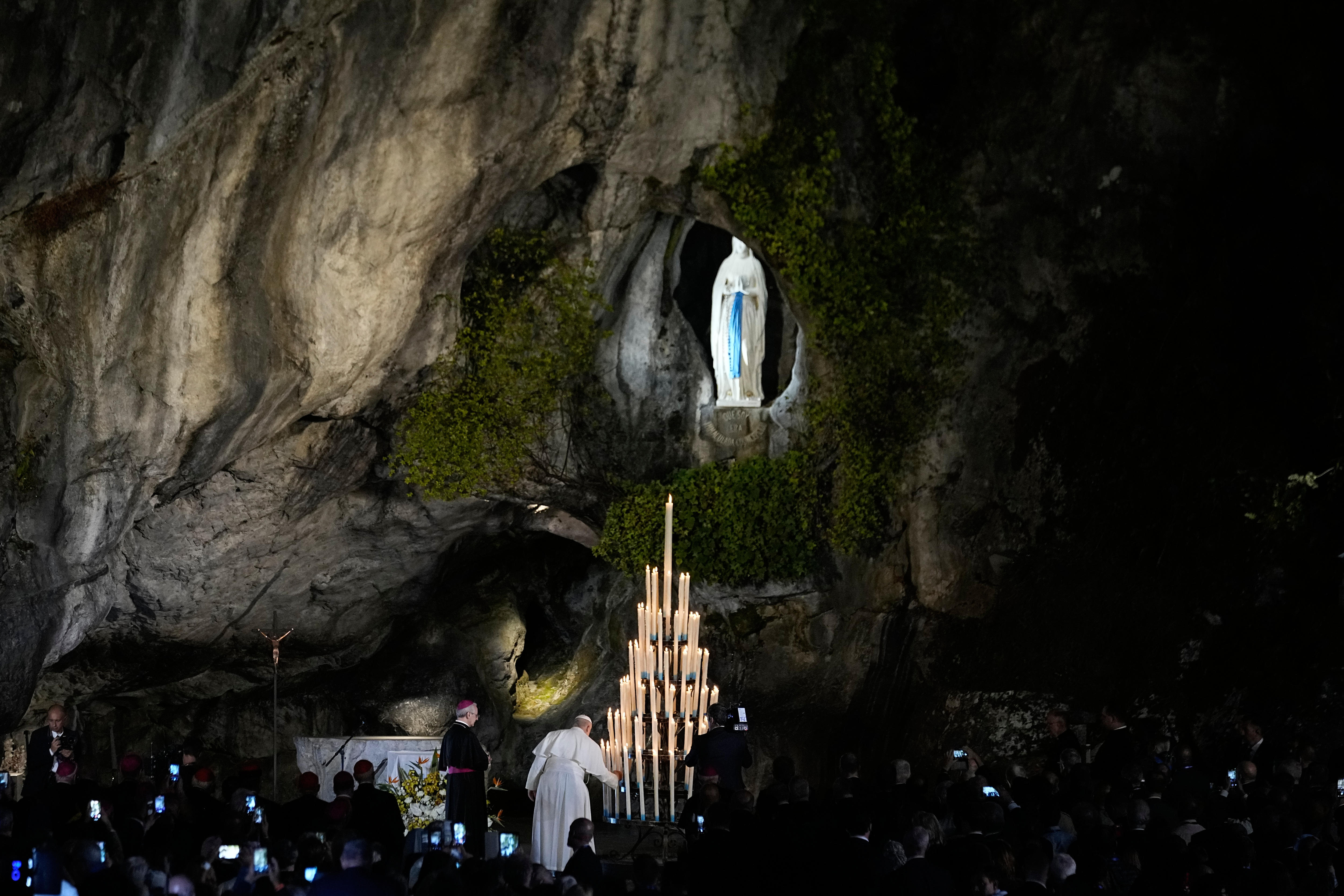 Pope Leo lights a candle on a large stand of candles inside a cave, a crowd looks on.