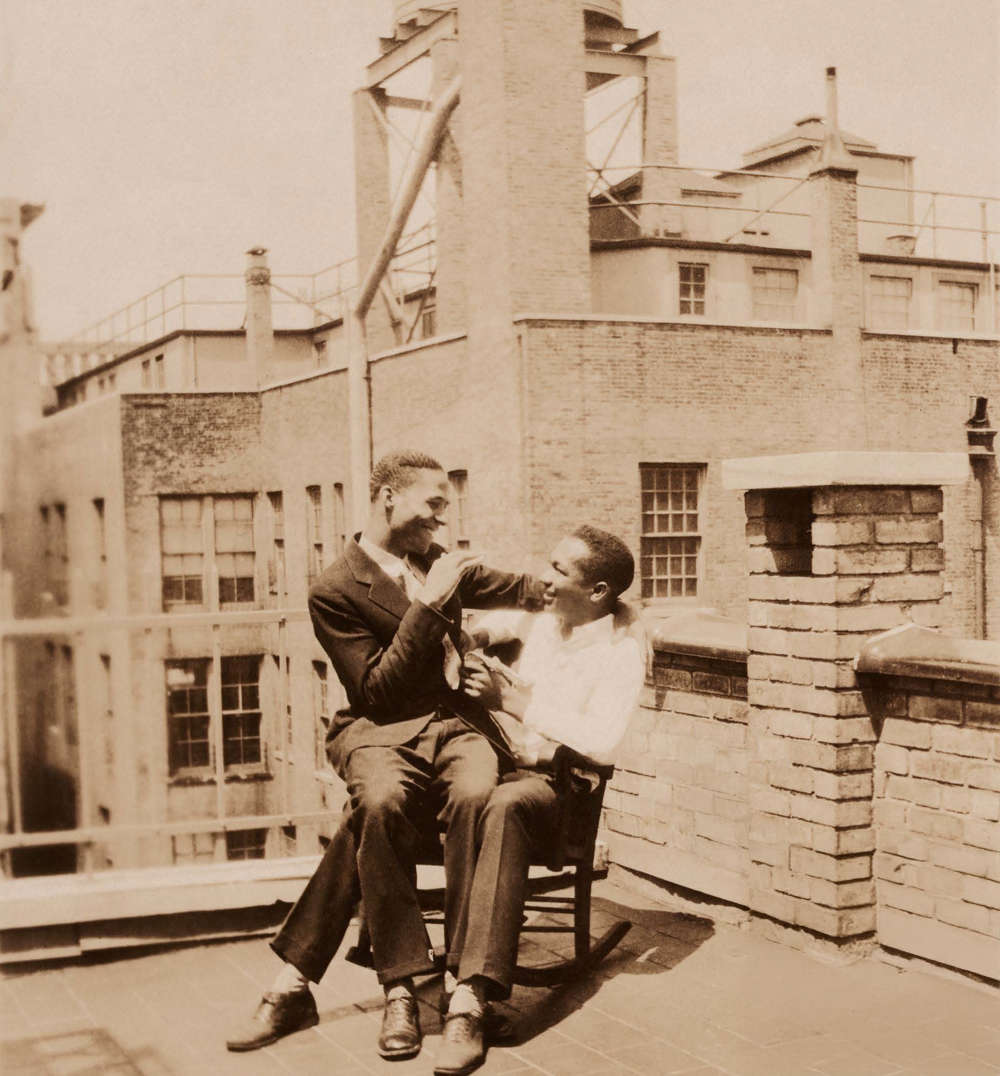 Un hombre se sienta en el regazo de otra persona en una silla en la azotea de un edificio.