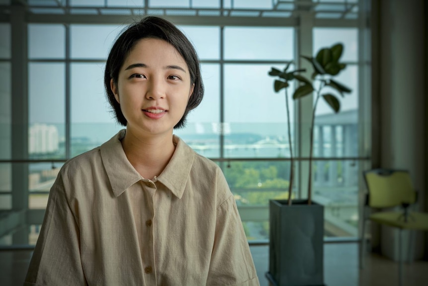 A young Korean woman in a red dress and yellow face mask sits at a desk 