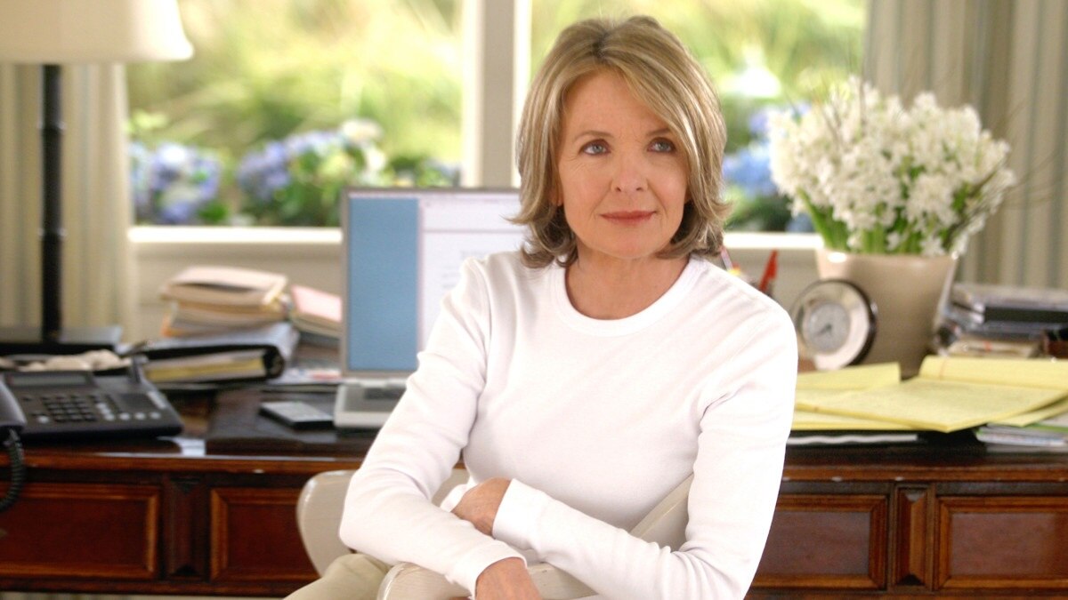 Diane Keaton sits with her arms cross on a chair at a desk in a brightly lit home.