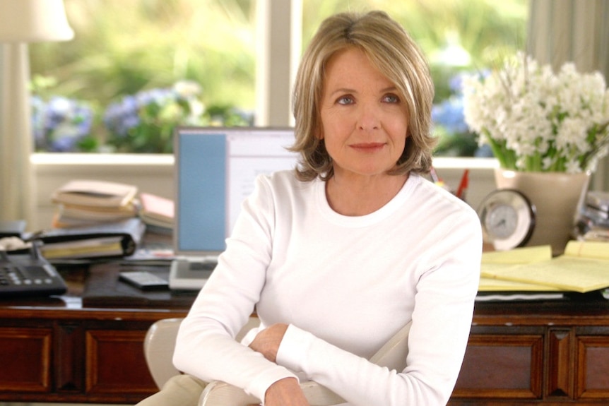 Diane Keaton sits with her arms cross on a chair at a desk in a brightly lit home.
