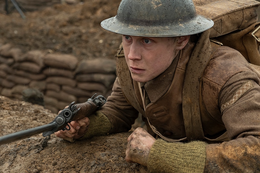 A man in world war one brown uniform, helmet and rifle stands at the edge of trench looking into distance on a rainy day.