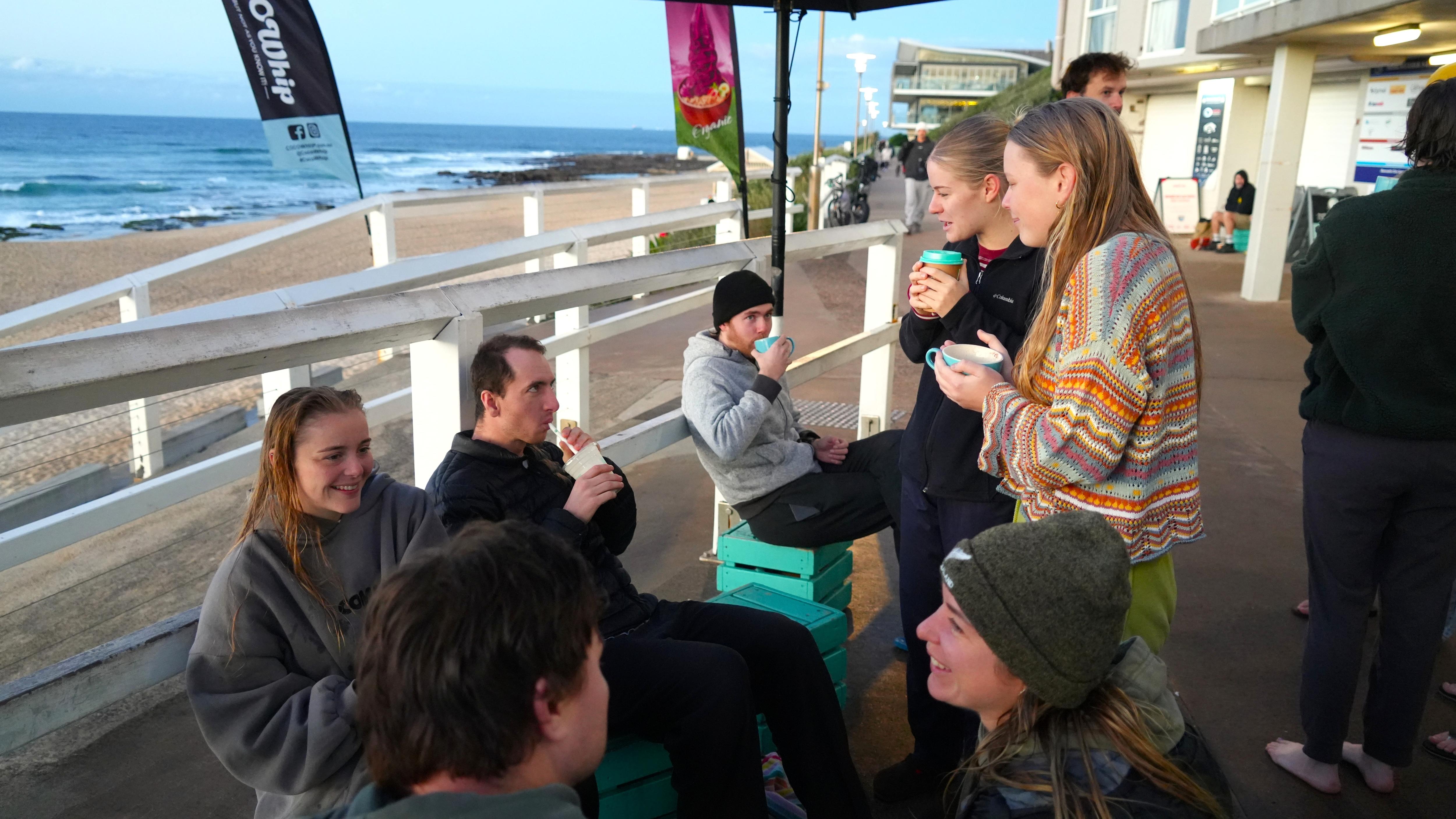 A group of people stand around drinking coffee near the beach.